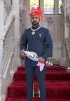 Embroidered deer logo coat suit in navy worn by a man holding a traditional weapon in an ornate setting
