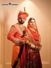 A groom in a vibrant orange men's sherwani angarkha paired with traditional accessories alongside the bride