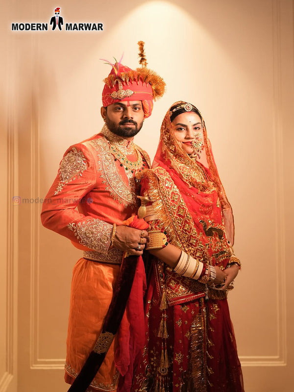 A groom in a vibrant orange men's sherwani angarkha paired with traditional accessories alongside the bride