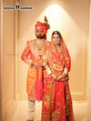 Men's sherwani angarkha in vibrant orange worn by a groom alongside a bride in traditional attire